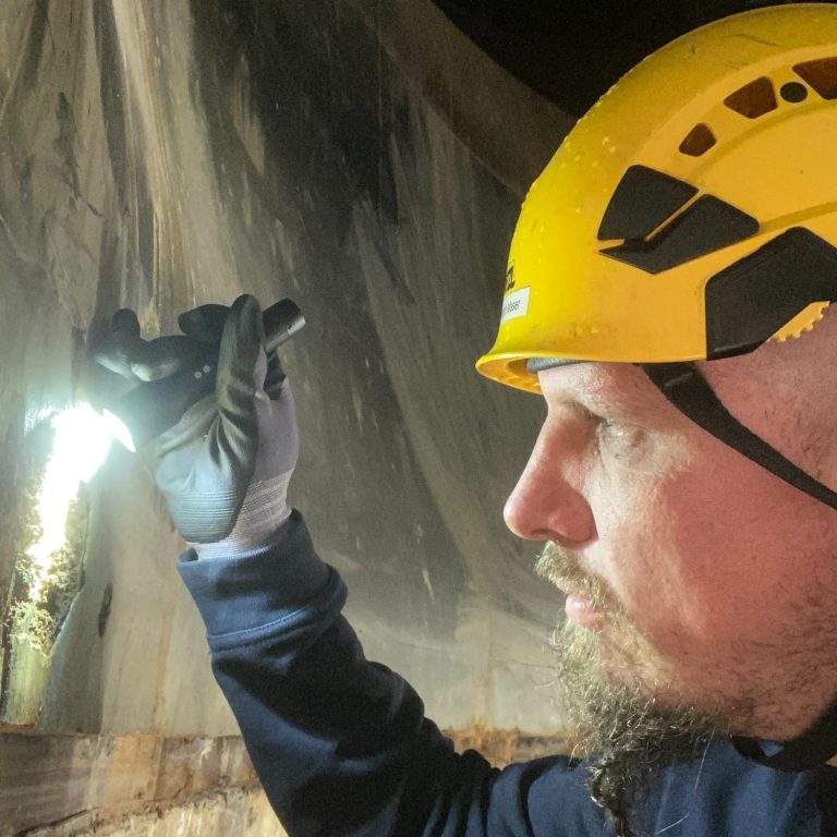 Mann mit Helm und Sicherheitsjacke, lächelt vor grünem Hintergrund.